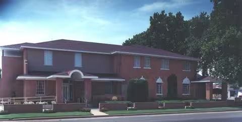 Brick two-story building with arched entry, windows, and landscaping on a sunny street.