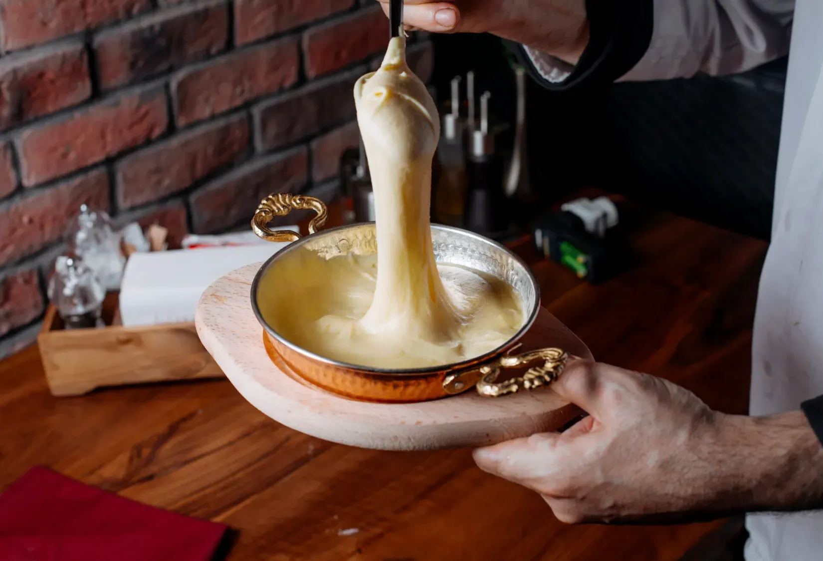 Mão de um chef segurando um recipiente de metal onde o queijo derretido do fondue é puxado para cima, mostrando sua textura cremosa durante o Festival de Fondue em Gramado, próximo ao Hotel Recanto da Serra.