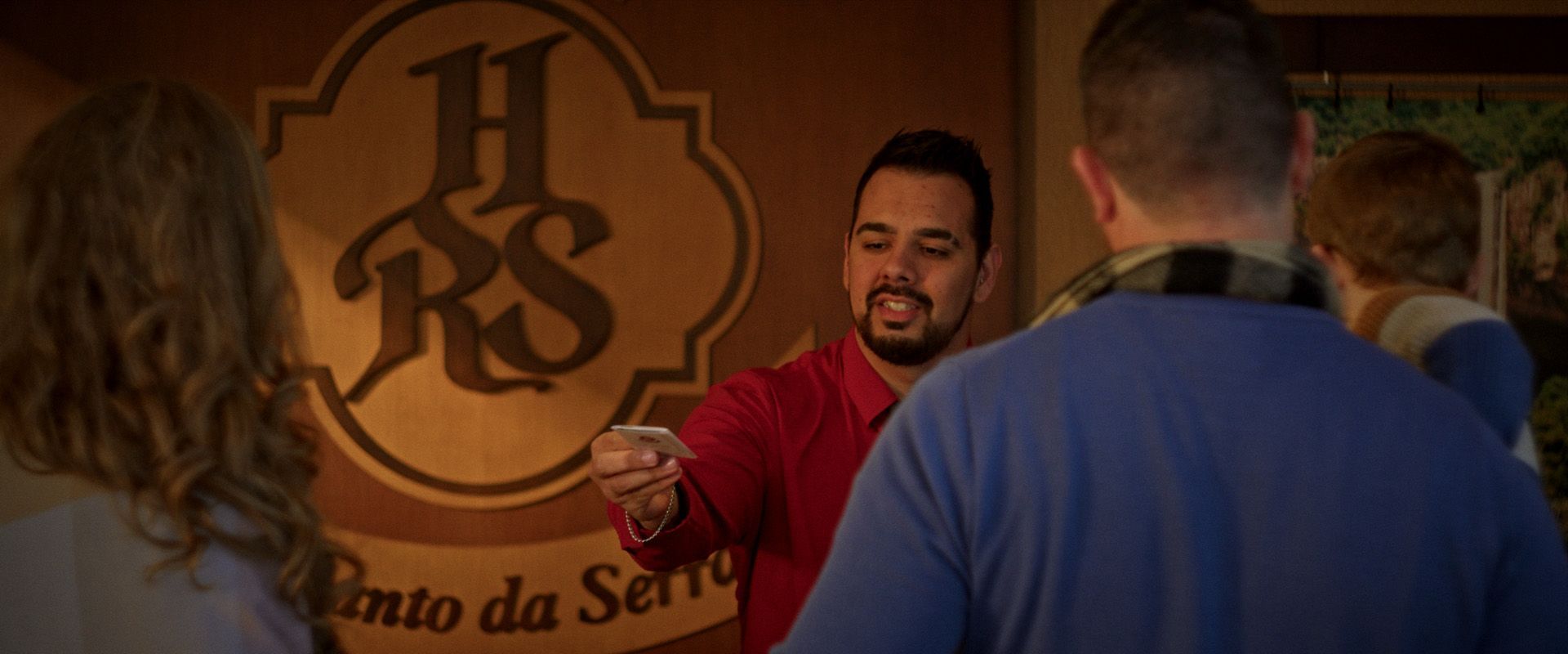 Homem de camisa vermelha distribuindo um panfleto em frente a uma parede com o logotipo “HRS” e o texto “Todo da Ser”.