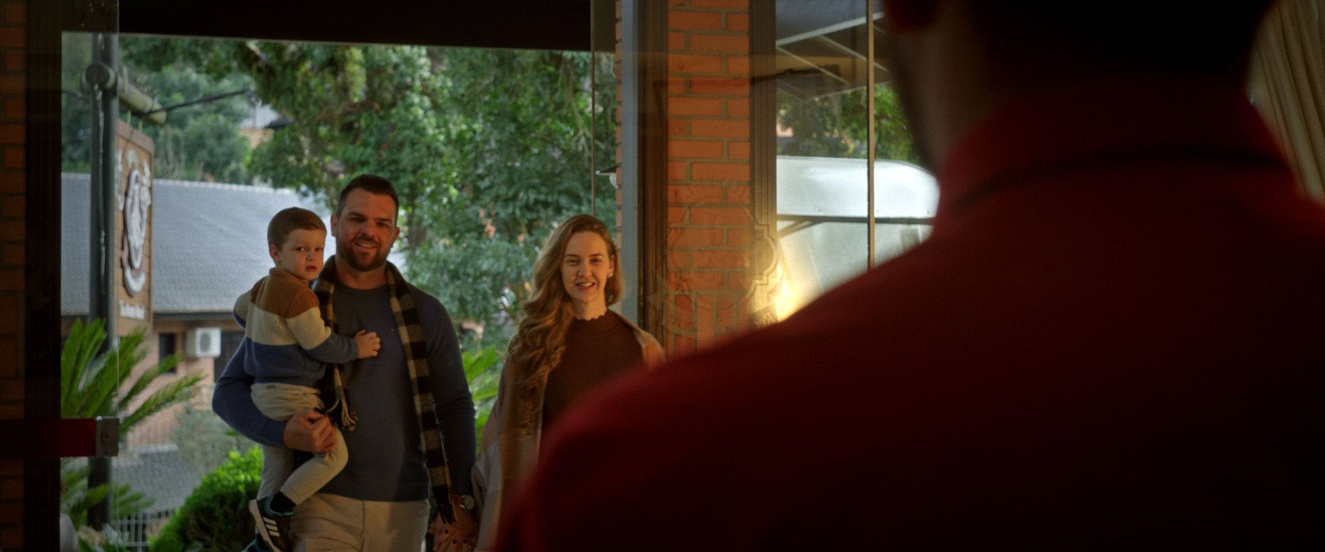 Família à porta de casa, com um homem segurando uma criança e uma mulher sorrindo lá dentro.
