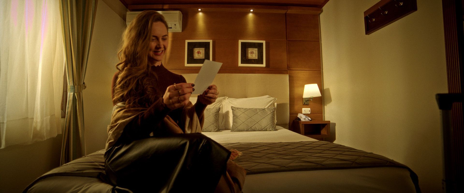 Mulher sentada na cama de um hotel lendo uma carta em um quarto com luz quente.