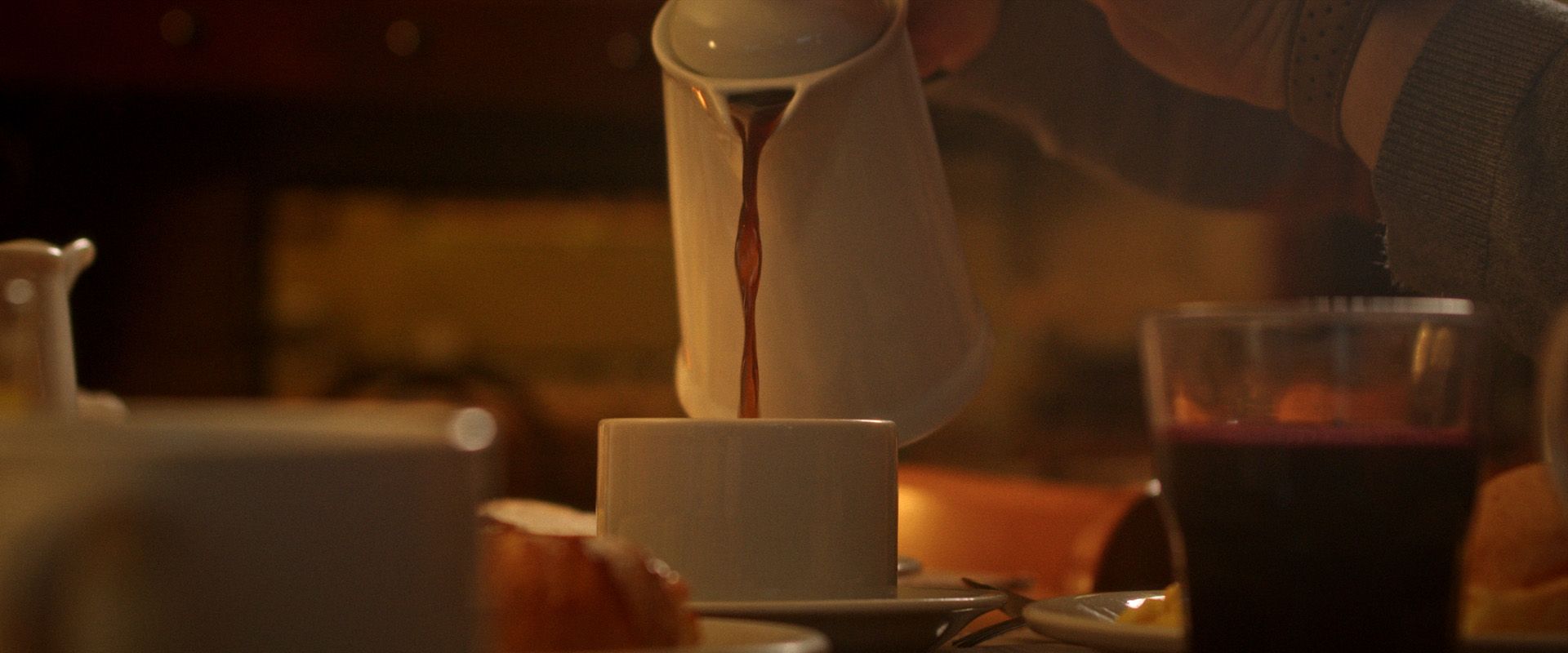 Mesa aconchegante e com luz baixa, com uma xícara e pires brancos, um bule de chá servindo e copos ao fundo.