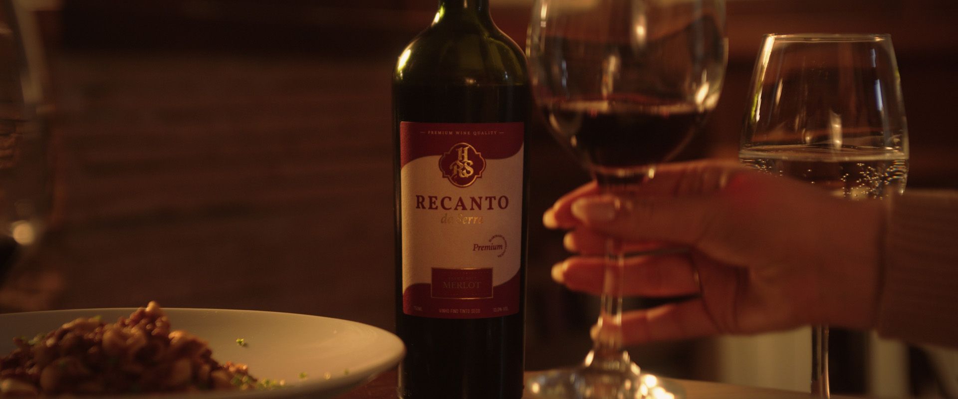 Mão segurando uma taça de vinho ao lado de uma garrafa de vinho em uma mesa de jantar com um prato de comida.