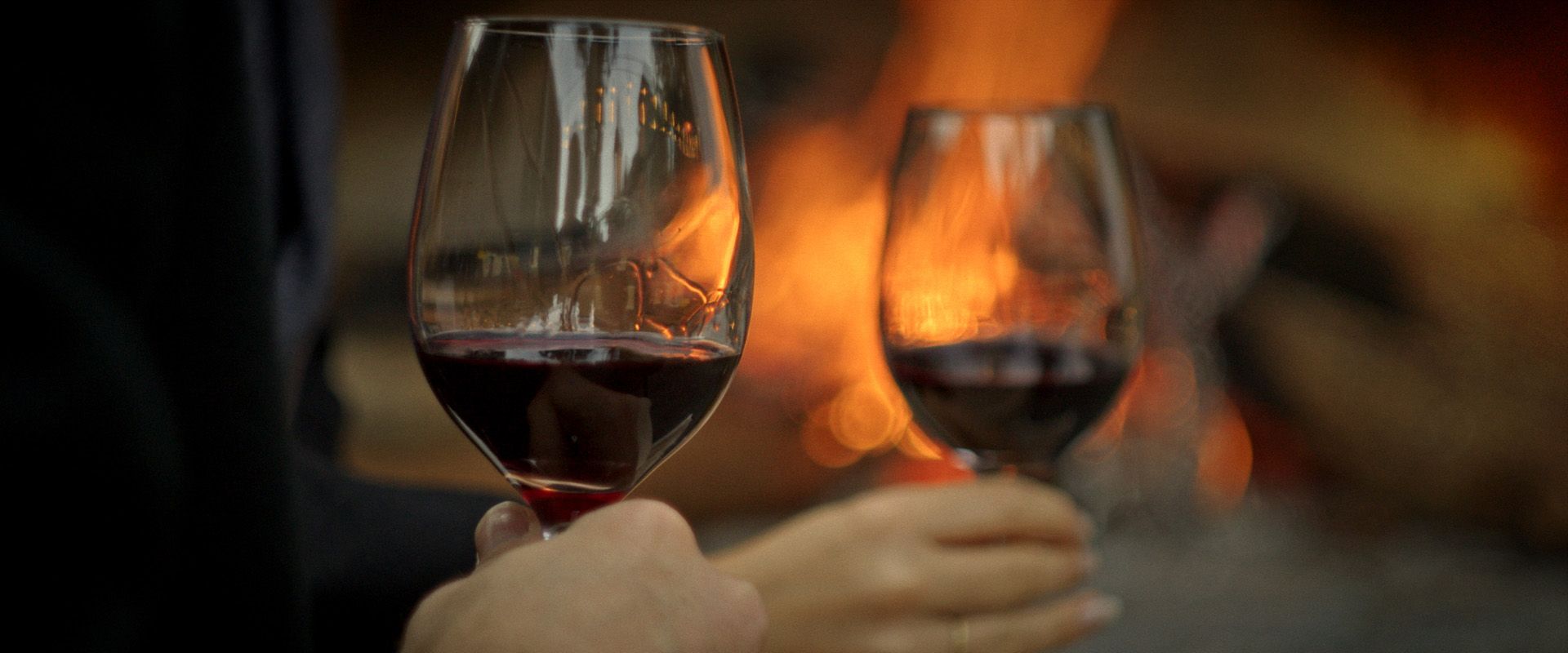 Duas mãos erguem taças de vinho tinto junto a uma lareira aconchegante.