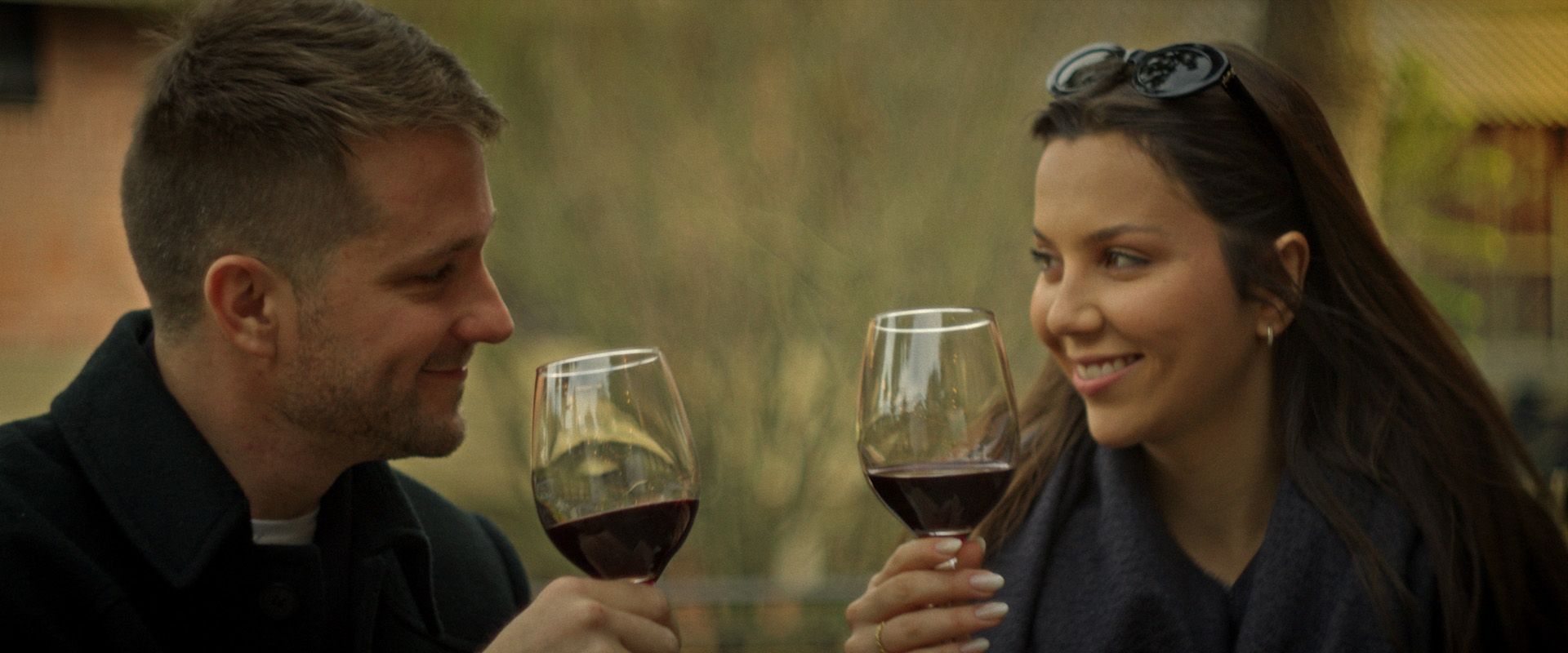 Duas pessoas brindando com taças de vinho tinto ao ar livre, sorrindo em um vinhedo.