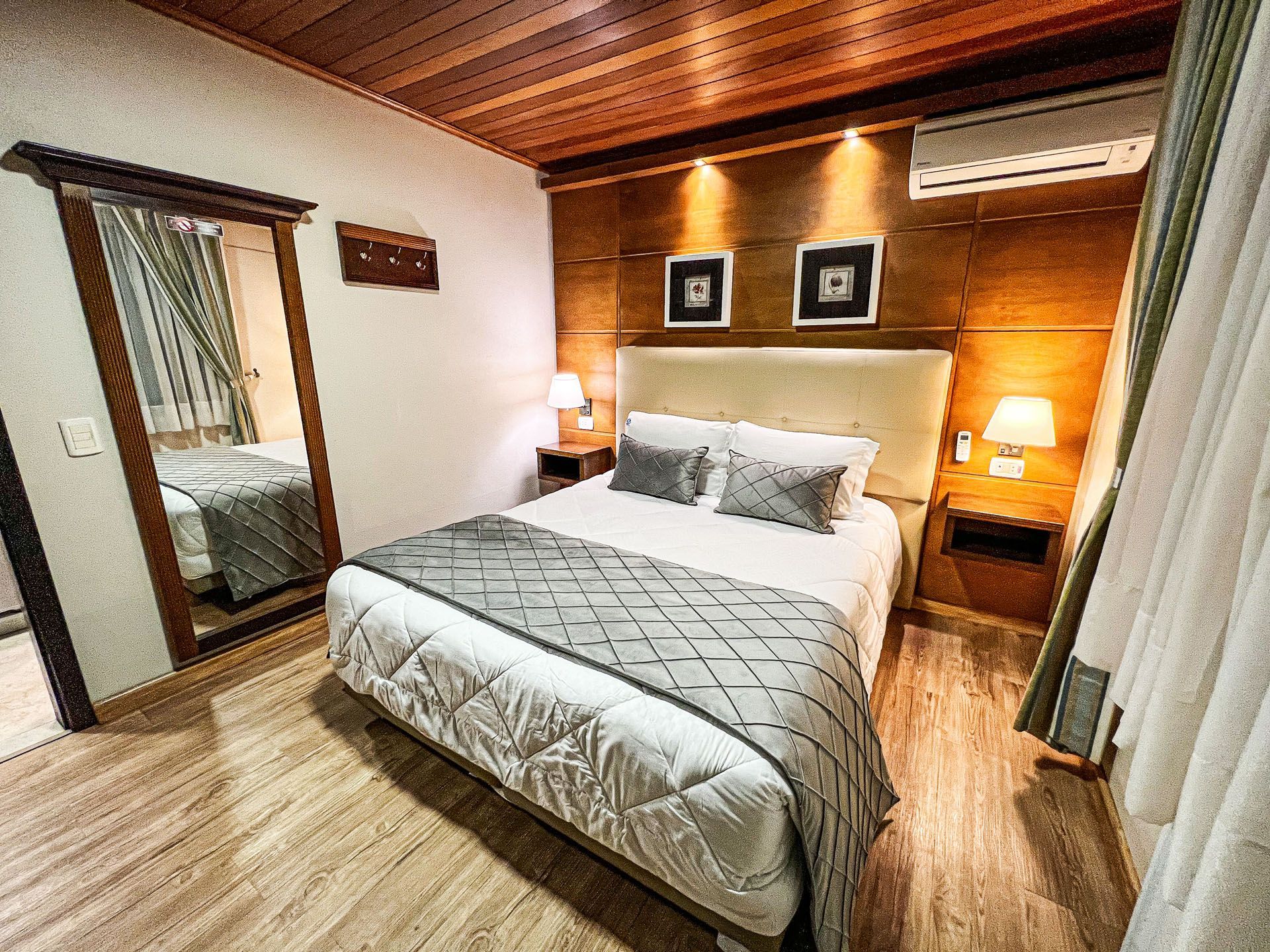 Quarto aconchegante com cama de casal, teto de madeira, obras de arte na parede, luminárias e portas de armário espelhadas.