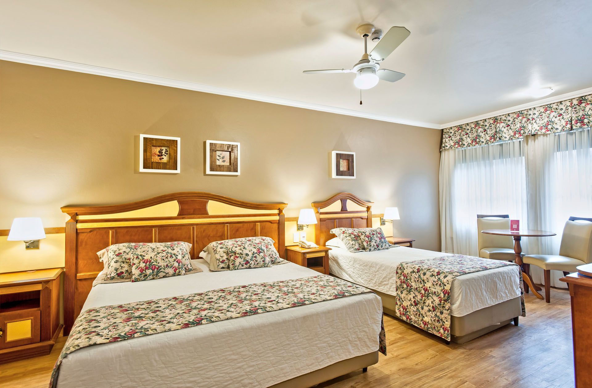 Quarto de hotel luminoso com duas camas floridas, luminárias e uma área de estar junto a grandes janelas.