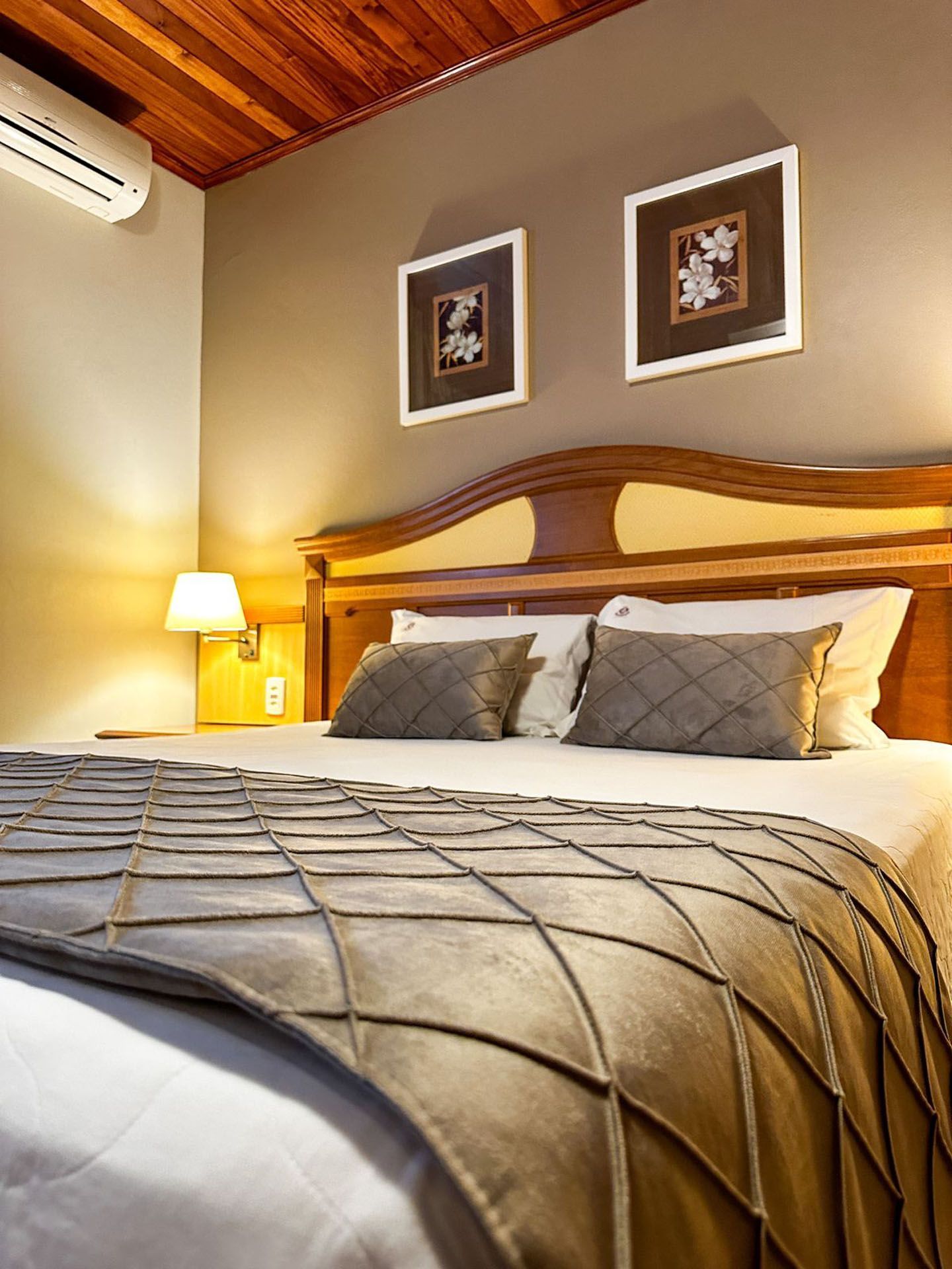 Quarto aconchegante com cabeceira de madeira, cama arrumada, luz quente de abajur e quadros na parede.