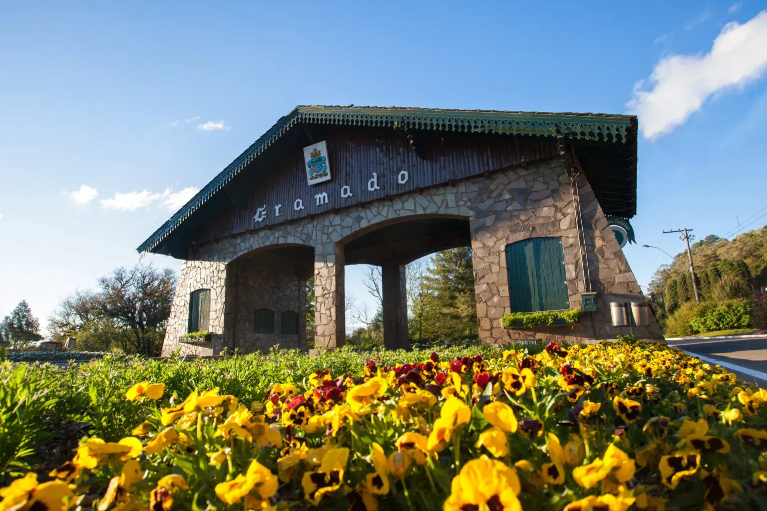 Foto em ângulo baixo do Pórtico de Gramado via Nova Petrópolis, construído em pedra e madeira com estilo bávaro. Em primeiro plano, um canteiro repleto de flores amarelas e escuras floresce sob um céu azul, destacando a beleza da primavera na Serra Gaúcha durante a baixa temporada.