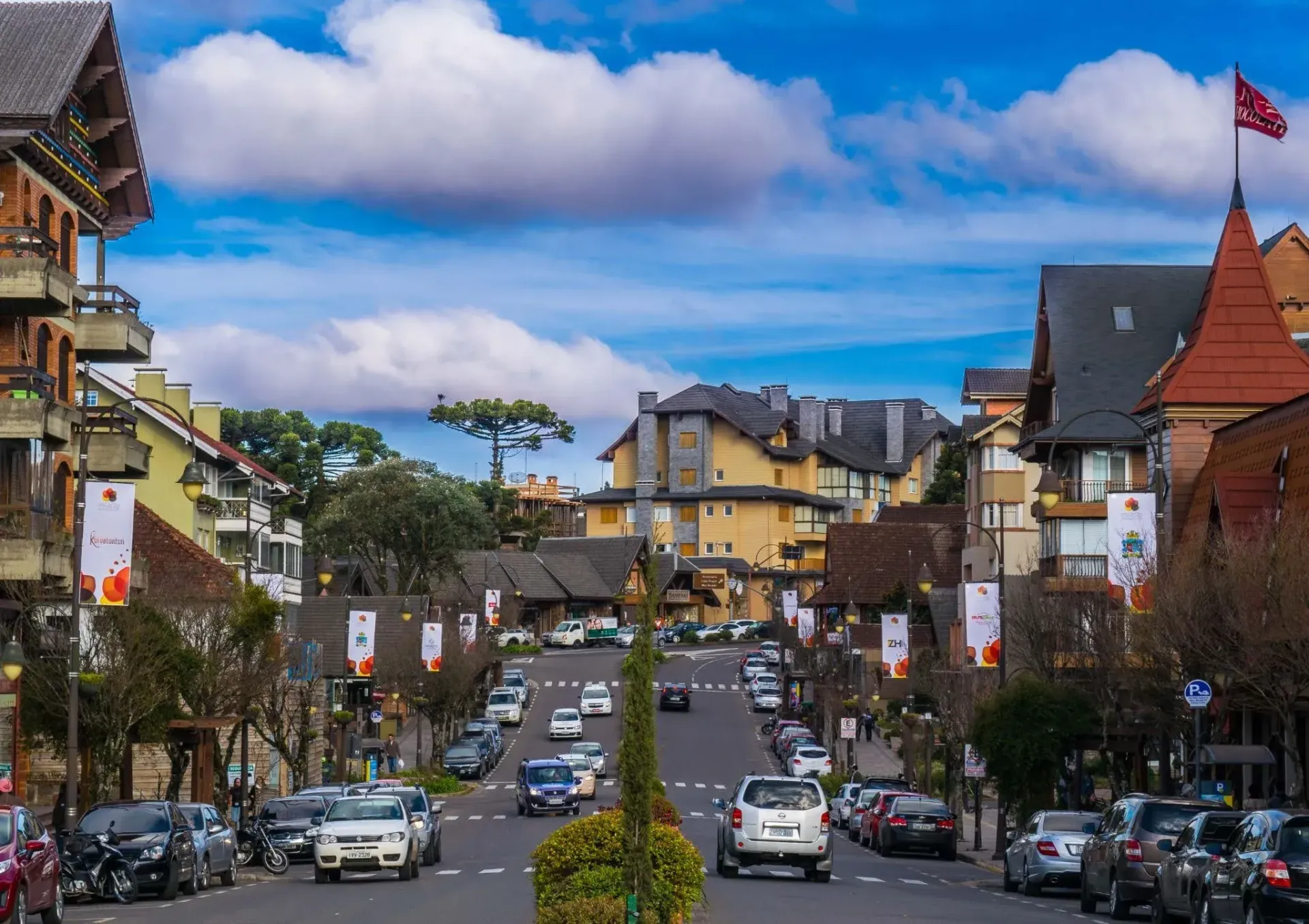 Vista central da Avenida Borges de Medeiros em Gramado, mostrando o fluxo tranquilo de carros entre prédios com arquitetura de estilo europeu e alpino. Banners decorativos enfeitam os postes e, ao fundo, árvores araucárias sobressaem sob um céu azul com grandes nuvens brancas, ilustrando o ritmo calmo da cidade.