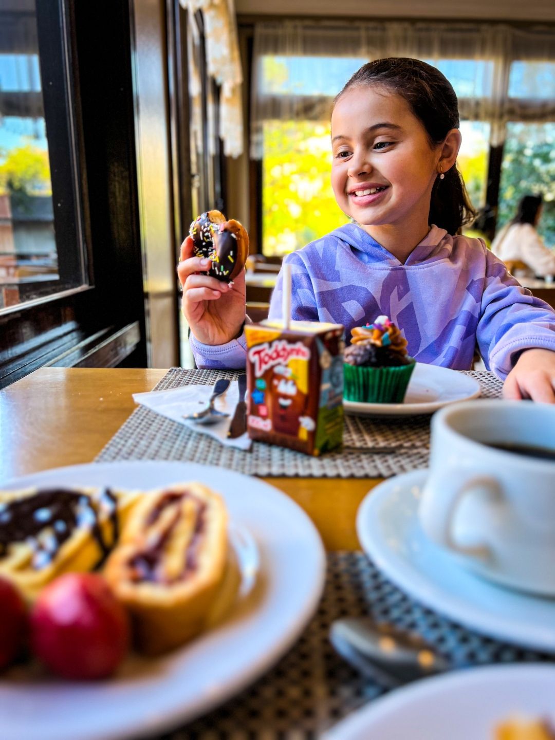 Pessoa sorridente segurando um donut em uma mesa de café com café e doces.