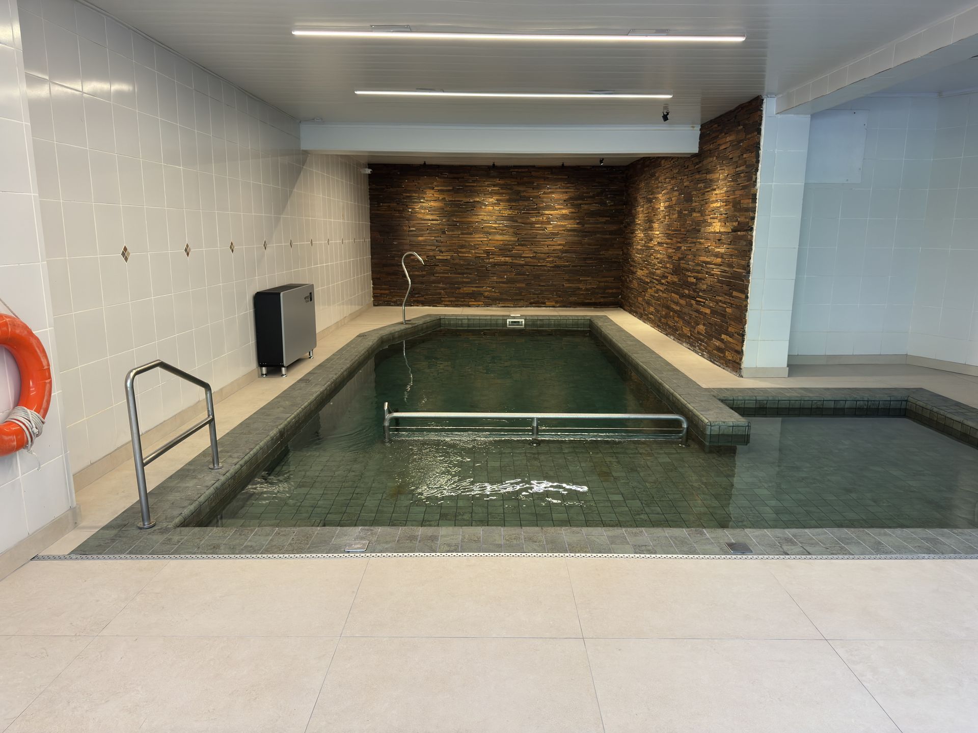 Piscina coberta com água cristalina, paredes revestidas com azulejos e uma pequena área de spa sob luzes brilhantes no teto.