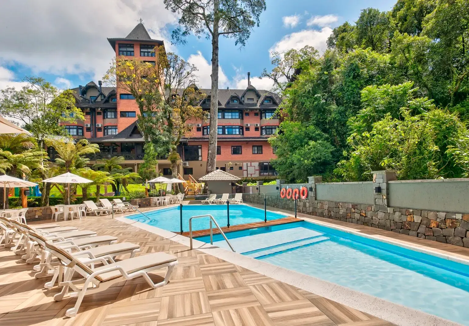Piscina em frente a um hotel com torre, rodeada por árvores e espreguiçadeiras.
