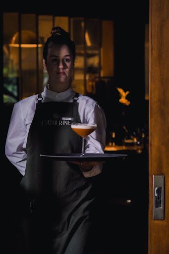 Garçom segurando um coquetel e uma bandeja na porta pouco iluminada de um restaurante.