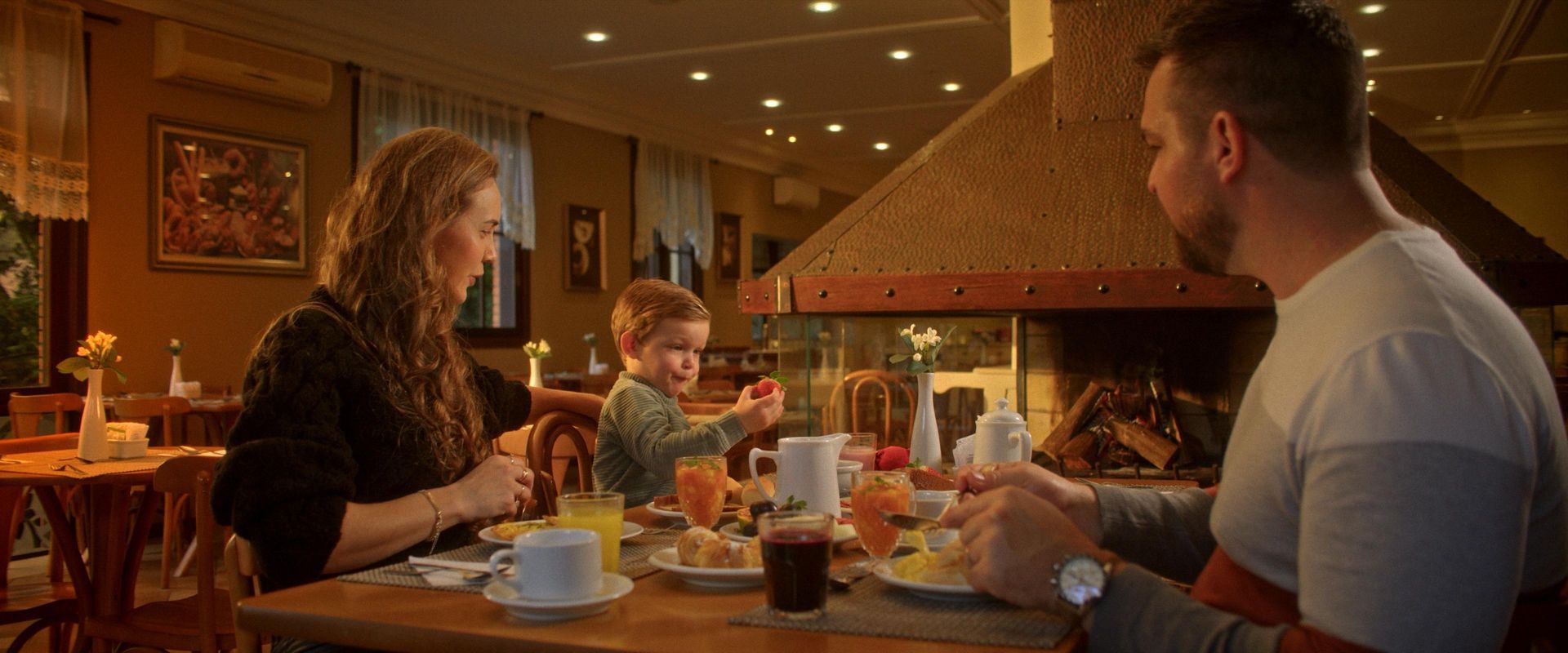Casal e criança tomando café da manhã colonial ao lado da lareira, no restaurante do Hotel Recanto da Serra, em Gramado.