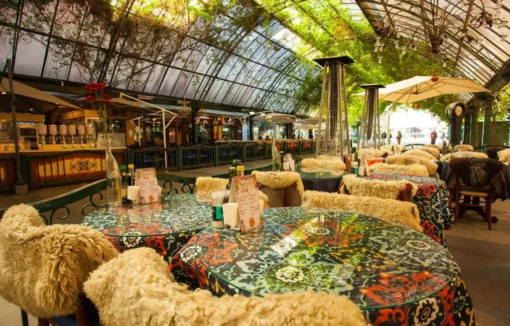 Restaurante ao ar livre com cadeiras cobertas de palha e mesas coloridas sob um dossel frondoso.
