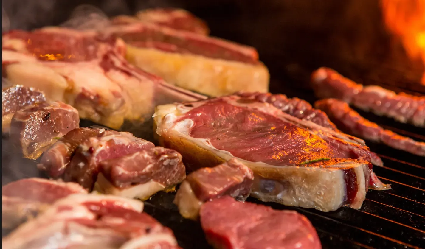 Cortes nobres de carne vermelha e linguiças sendo assados na grelha sobre chamas vivas, representando a tradição do churrasco gaúcho em Gramado, próximo ao Hotel Recanto da Serra.