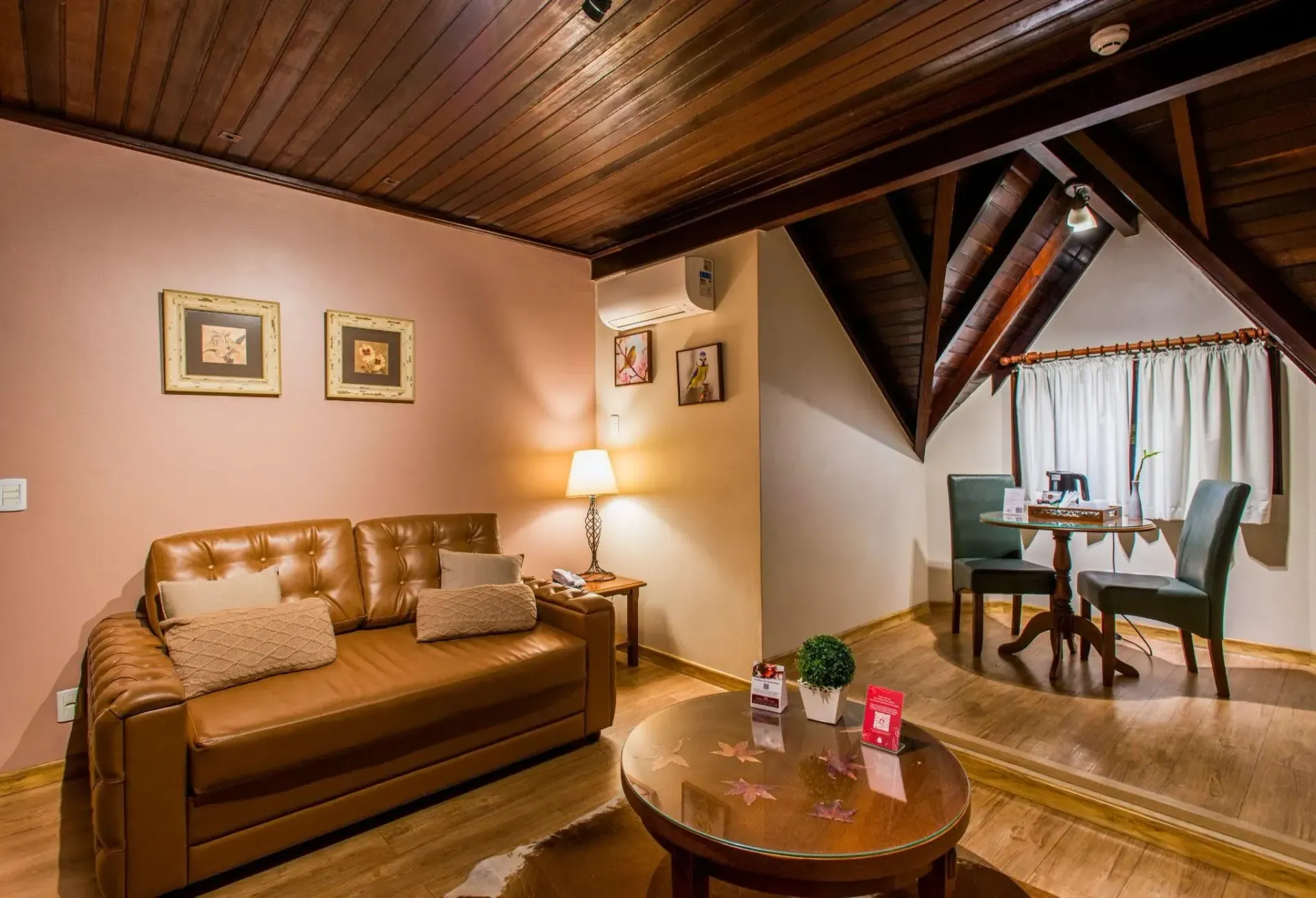 Interior aconchegante de uma acomodação do hotel, com sofá de couro marrom, mesa redonda com cadeiras, piso de madeira e teto inclinado com vigas aparentes.