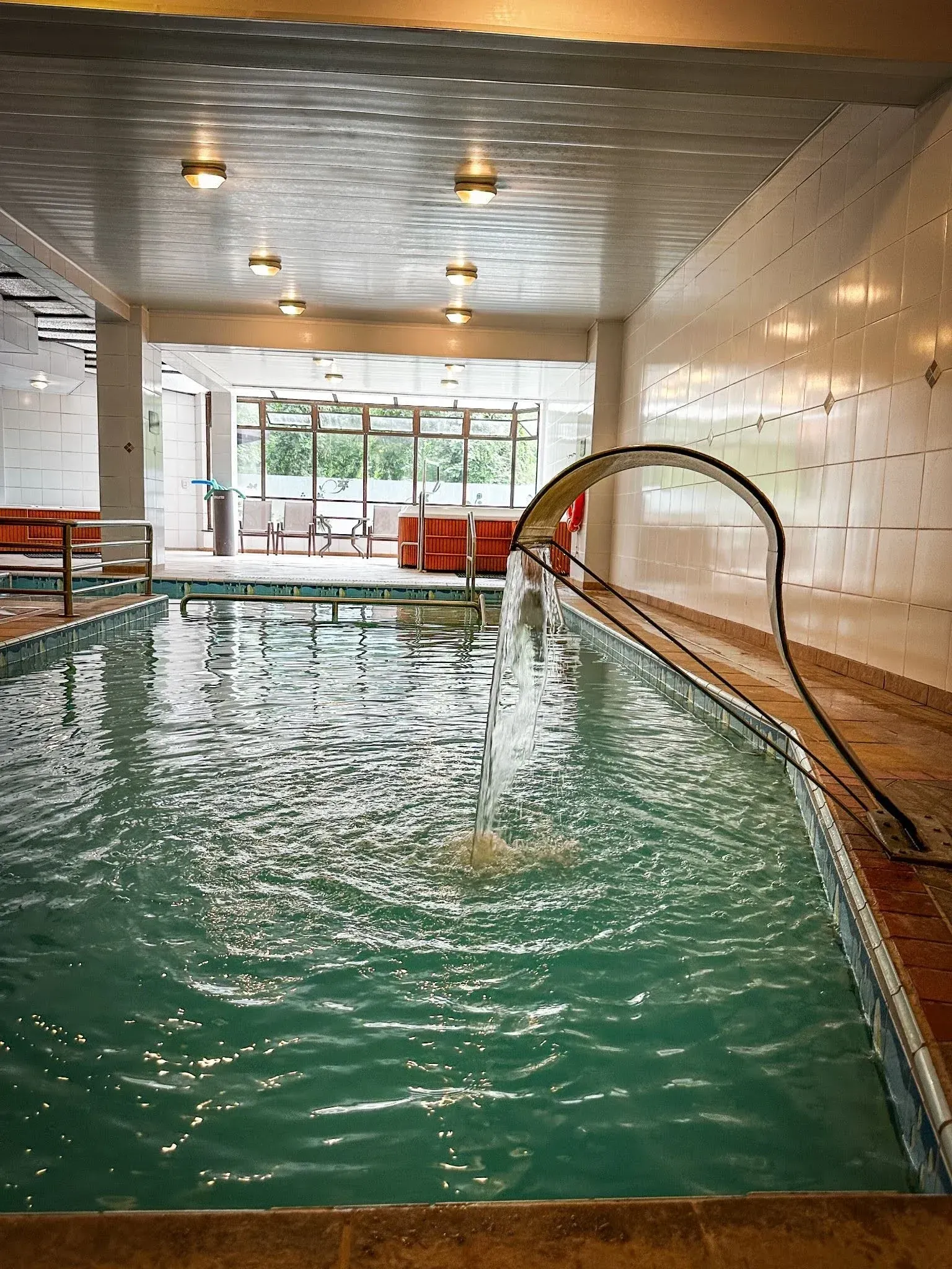 Piscina coberta e aquecida do Hotel Recanto da Serra, destacando uma cascata d'água caindo na piscina e espreguiçadeiras ao fundo próximas às janelas.