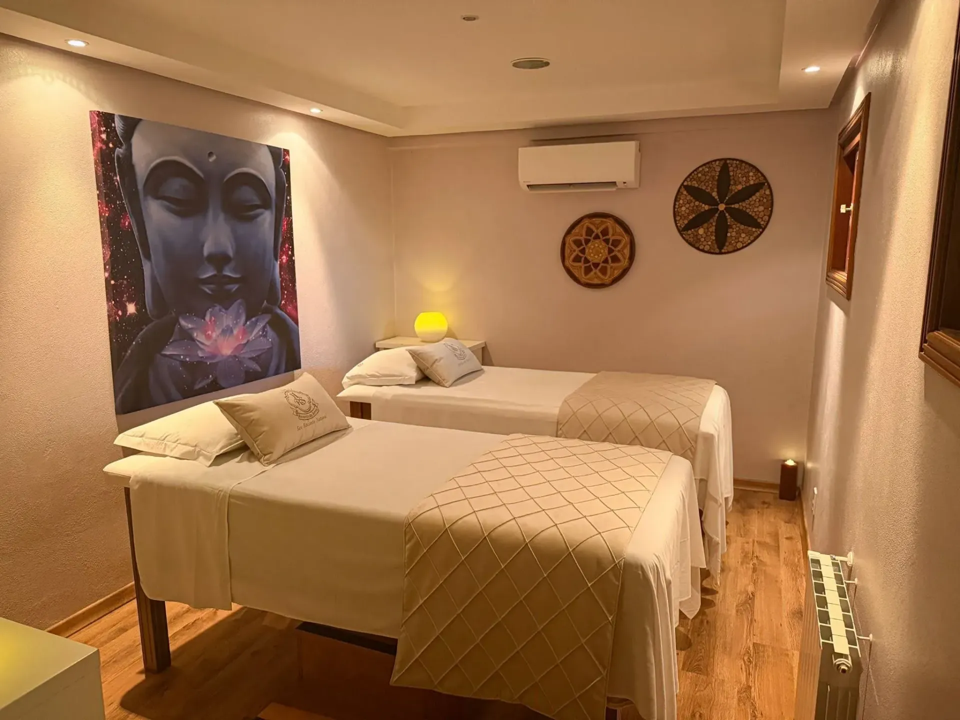 Sala do SPA Recanto da Serra decorada em tons suaves, contendo duas macas de massagem arrumadas lado a lado e um quadro na parede com a imagem de Buda segurando uma flor de lótus.