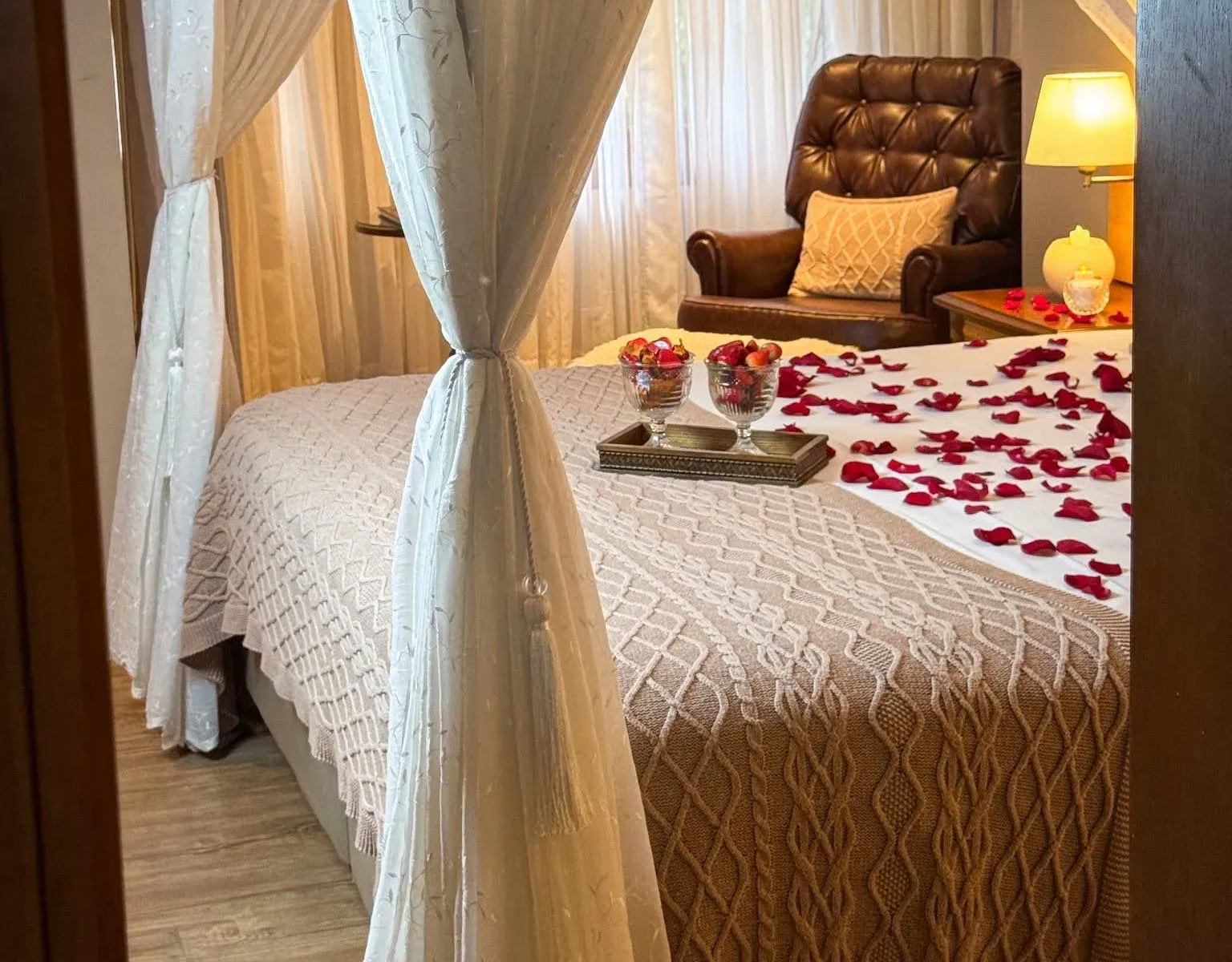 Suíte aconchegante do Hotel Recanto da Serra com cama de dosseis, iluminação suave, pétalas de rosas espalhadas e taças com morangos e chocolates do Pacote Speciale.