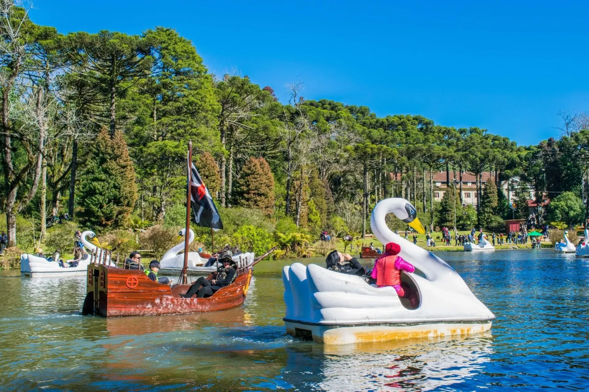 Pedalinhos em formato de cisne branco e um em formato de caravela navegam pelas águas calmas do Lago Negro em Gramado, cercado por árvores araucárias e turistas na margem, sob um céu azul limpo em um dia de sol.