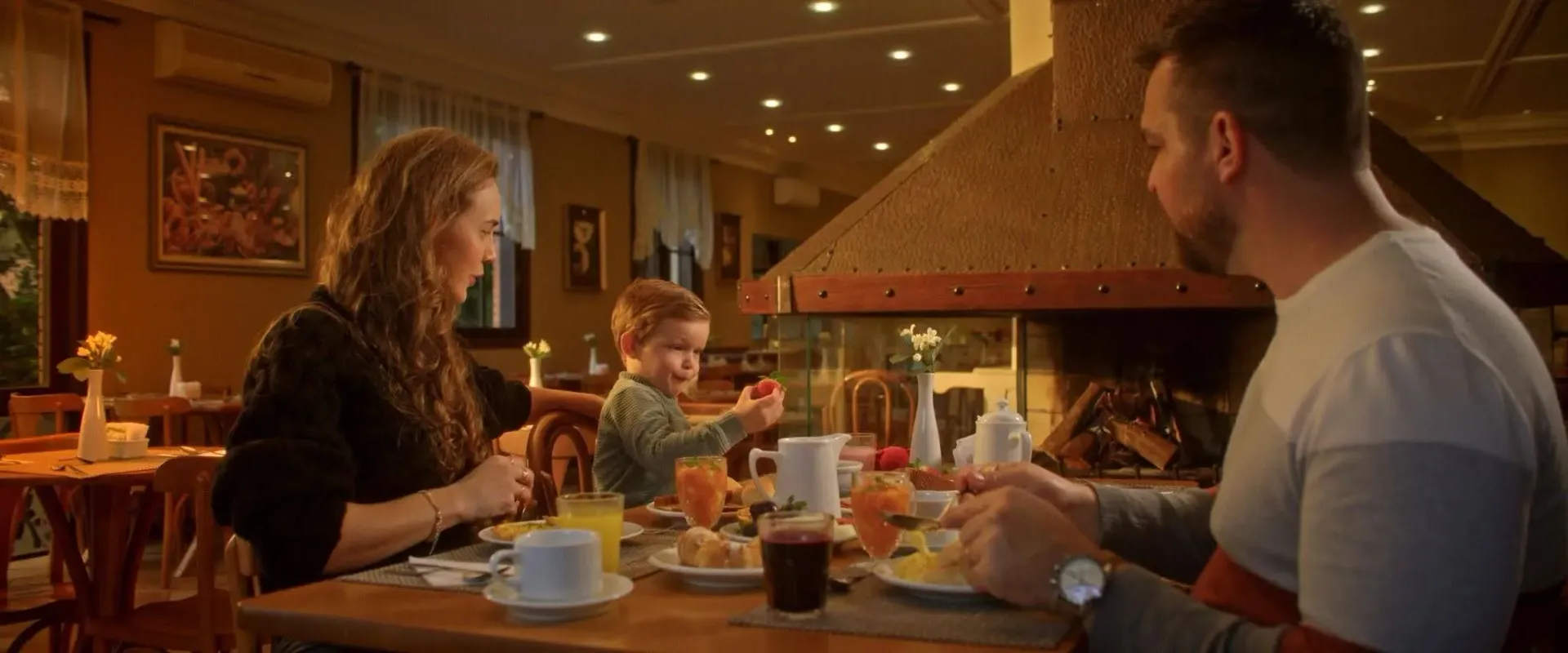 Uma família reunida aproveitando a gastronomia das férias de inverno na Serra Gaúcha dentro do Hotel Recanto da Serra. À mesa, uma mulher, um homem e um menino pequeno tomam café da manhã em um ambiente acolhedor, iluminado em tons quentes, com uma grande lareira acesa ao fundo.