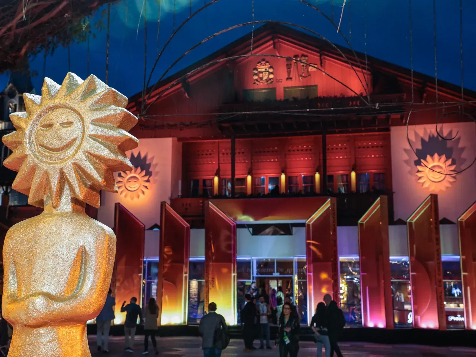 Uma fotografia noturna do Palácio dos Festivais iluminado em Gramado, com um troféu dourado em forma de sol em primeiro plano. Pessoas caminham em frente à entrada do local onde acontece o Festival de Cinema de Gramado.