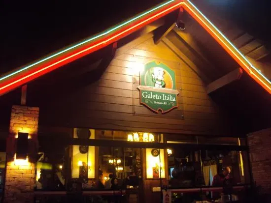 Fachada noturna do restaurante Galeto Itália. A estrutura de madeira sob um telhado triangular é iluminada por filetes de neon nas cores verde, vermelho e amarelo ao longo das bordas do teto. No centro da parede de madeira, há uma placa iluminada com o logotipo do restaurante, apresentando um desenho de chef de cozinha e o nome
