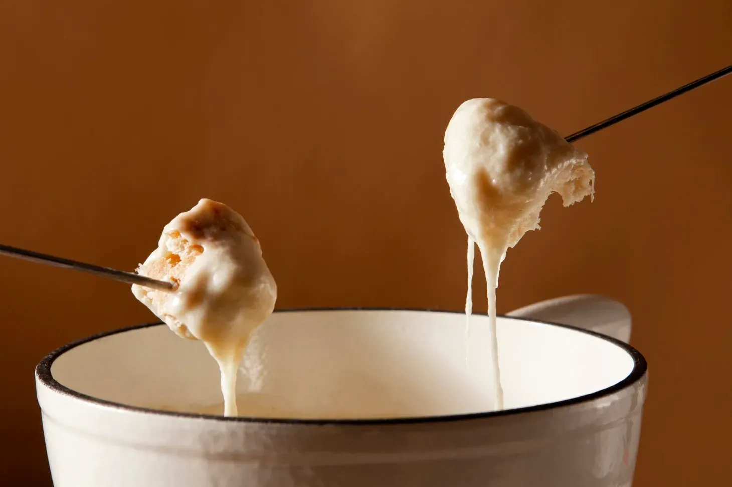 Dois pedaços de pão cobertos com queijo derretido escorrendo, espetados em garfos de fondue sobre uma panela de cerâmica branca, representando a tradicional sequência de fondue na gastronomia de Gramado.