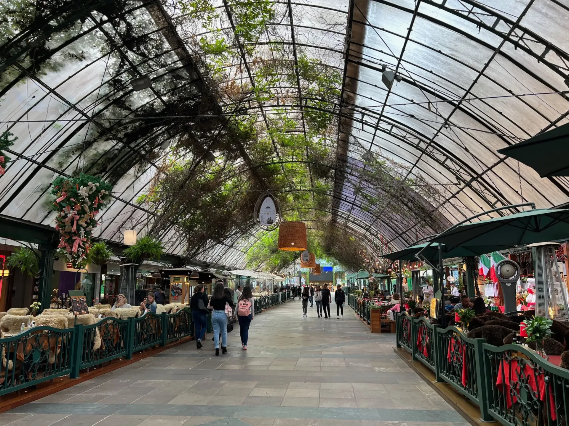 Galeria coberta com teto de vidro e trepadeiras na Rua Coberta em Gramado, com restaurantes e visitantes caminhando.