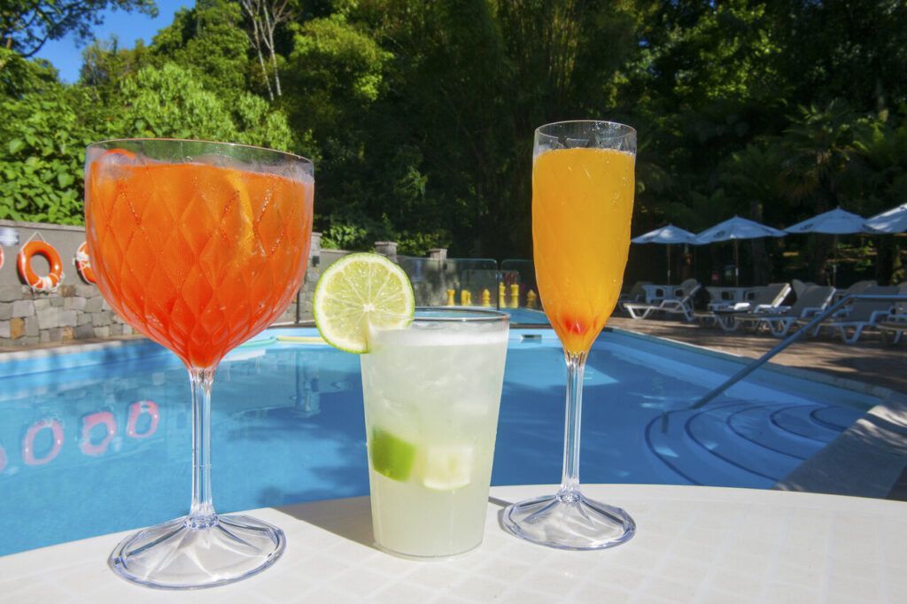 Três coquetéis coloridos à beira da piscina em um terraço ensolarado, com árvores e espreguiçadeiras ao fundo.