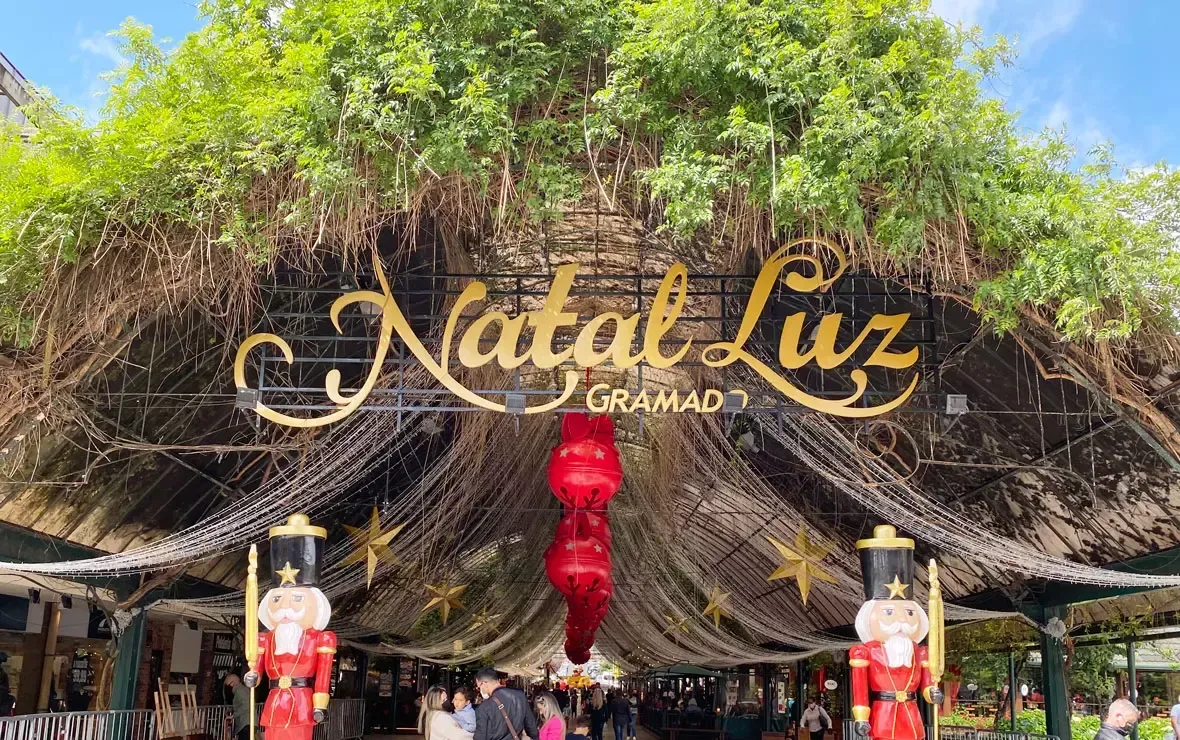 Placa de entrada com a inscrição “Natal Luz Gramado” e decorações festivas em vermelho sob um telhado de palha.