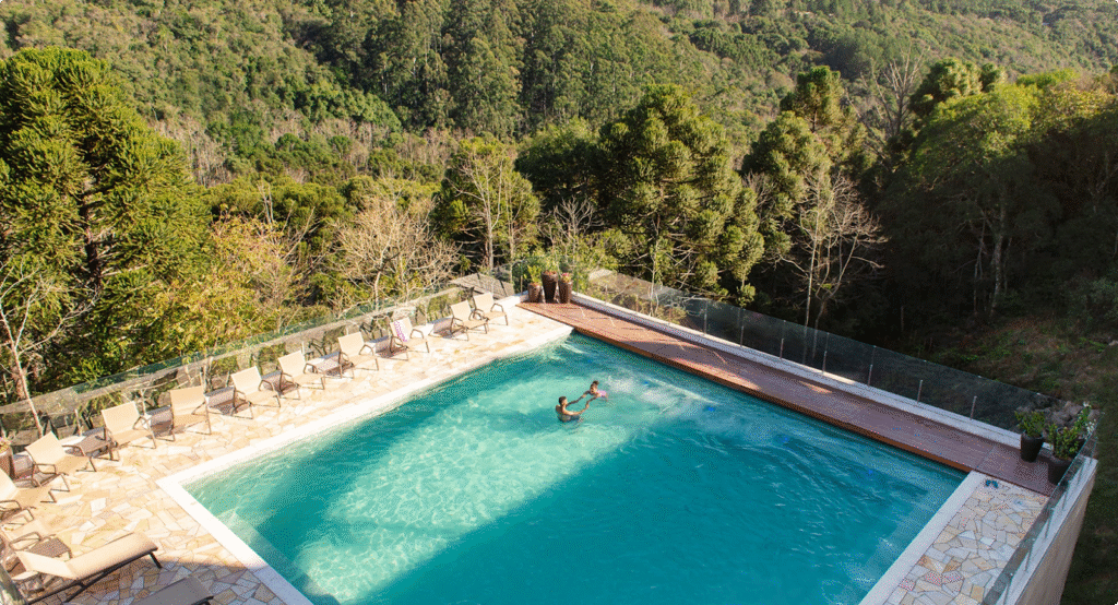 Piscina externa turquesa ao lado de uma encosta arborizada, com um nadador e espreguiçadeiras na borda.