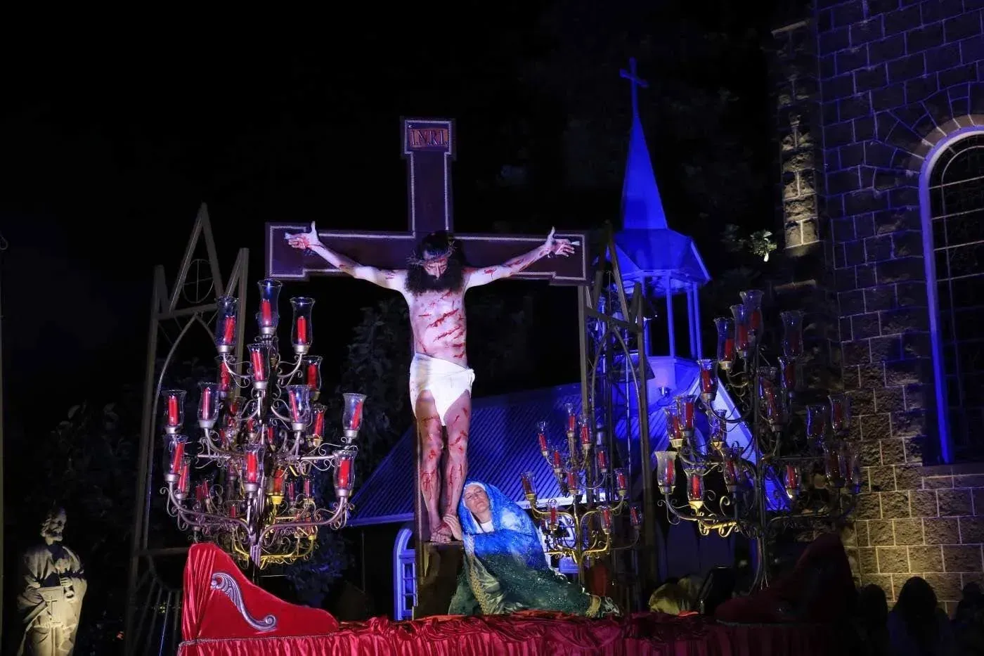 Cena da crucificação com um crucifixo aceso e uma estátua de Jesus em um palco, rodeados por velas e luzes roxo-azuladas.