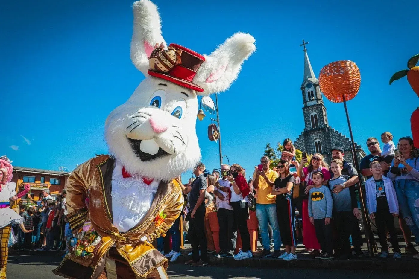 Mascote de coelho fantasiado com roupa de desfile vermelha e dourada, com multidão animada e torre da igreja ao fundo.