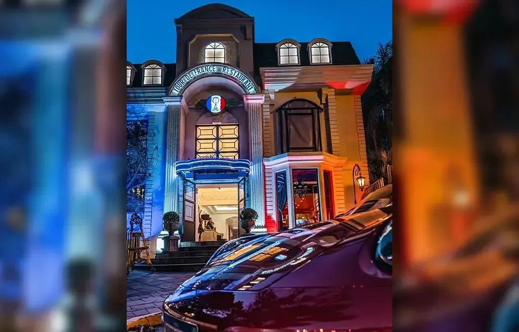 Fachada noturna impressionante do Tour de France Gramado, um restaurante que transporta os clientes para a Belle Époque. A foto noturna captura a elegância da arquitetura do edifício, com iluminação em tons de azul e vermelho que ressaltam os detalhes e o letreiro luminoso com o nome do restaurante e o icônico emblema da bandeira francesa. A entrada sofisticada e o ambiente refinado são visíveis, prometendo jantares românticos e celebrações especiais embaladas por pratos clássicos da culinária francesa com toques criativos. No primeiro plano, o detalhe refletido de um carro luxuoso completa a atmosfera de sofisticação.