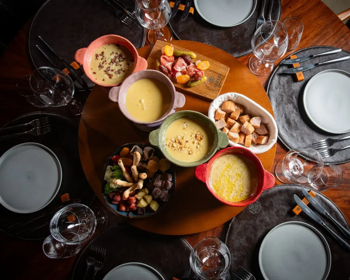 Vista aérea de uma mesa de jantar em madeira escura, posta para quatro pessoas, no requintado restaurante Catherine Gramado. A mesa apresenta uma sequência de fondue vibrante, com quatro panelas de cerâmica colorida contendo queijo derretido, acompanhadas por cestas de pães, uma tábua de frios, frutas secas, picles e diversas tigelas com acompanhamentos frescos e coloridos, como vegetais, batatas e frios. Taças de vinho vazias e pratos brancos minimalistas completam o cenário, prometendo uma experiência gastronômica autêntica e inesquecível, típica da Serra Gaúcha.