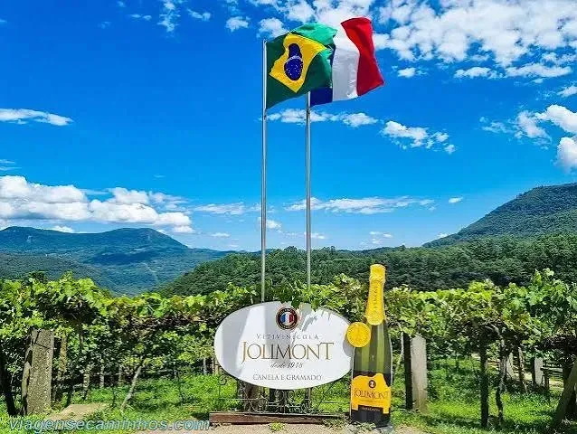 Placa de boas-vindas da Vitivinícola Jolimont em Canela com garrafa de espumante decorativa e bandeiras do Brasil e da França hasteadas, com vista para os parreirais e o vale de morros ao fundo.
