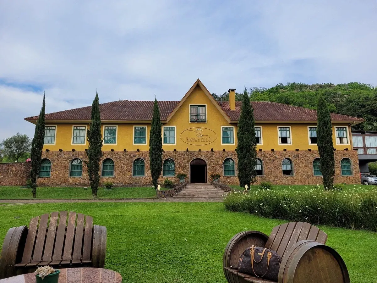 Vista frontal do imponente casarão amarelo e em pedras da Vinícola Ravanello em Gramado, com gramado verde bem cuidado, pinheiros e poltronas feitas com barris de vinho no jardim.