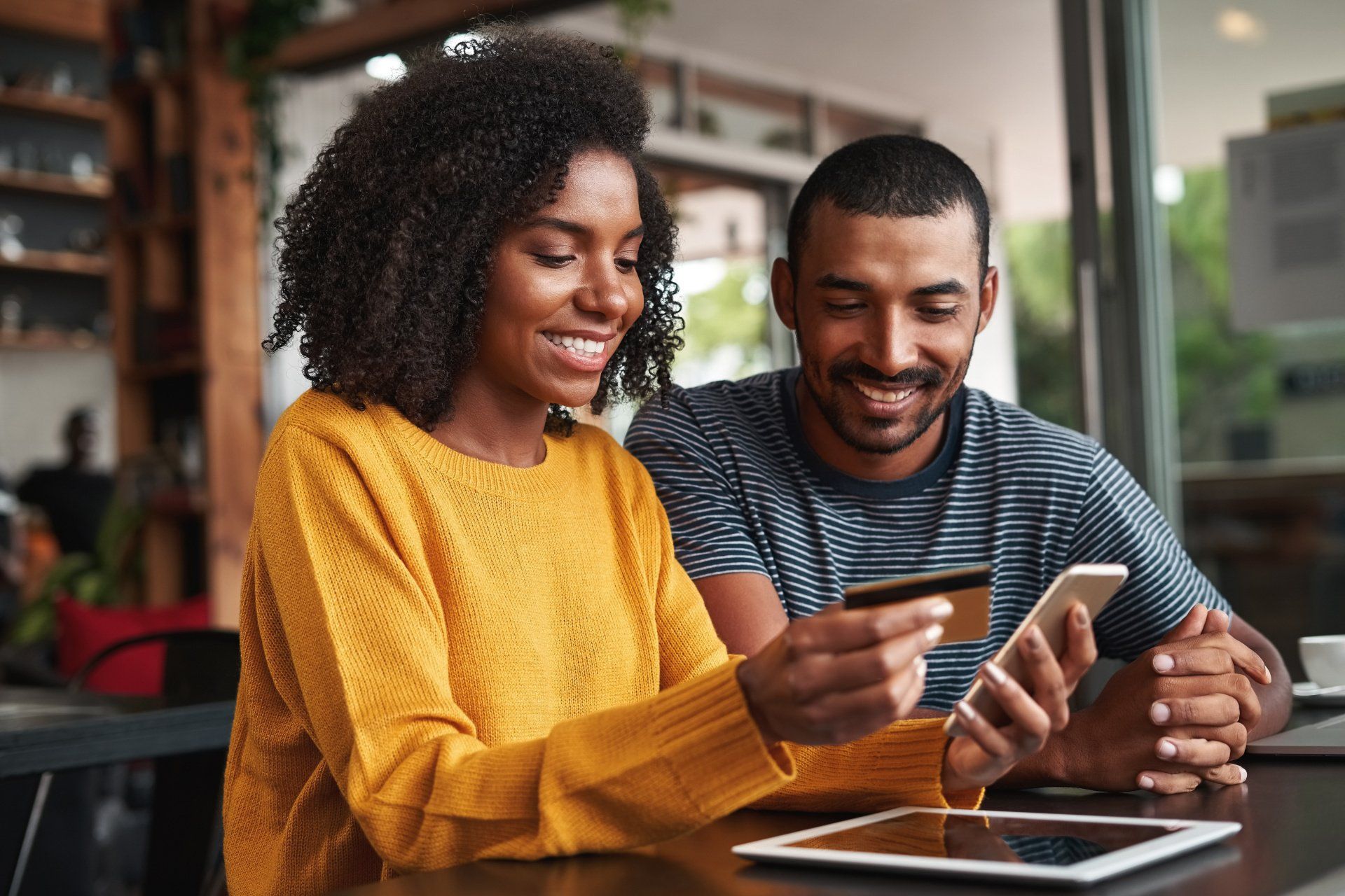 Woman holding a credit card and phone, man looking on, seated at a table.