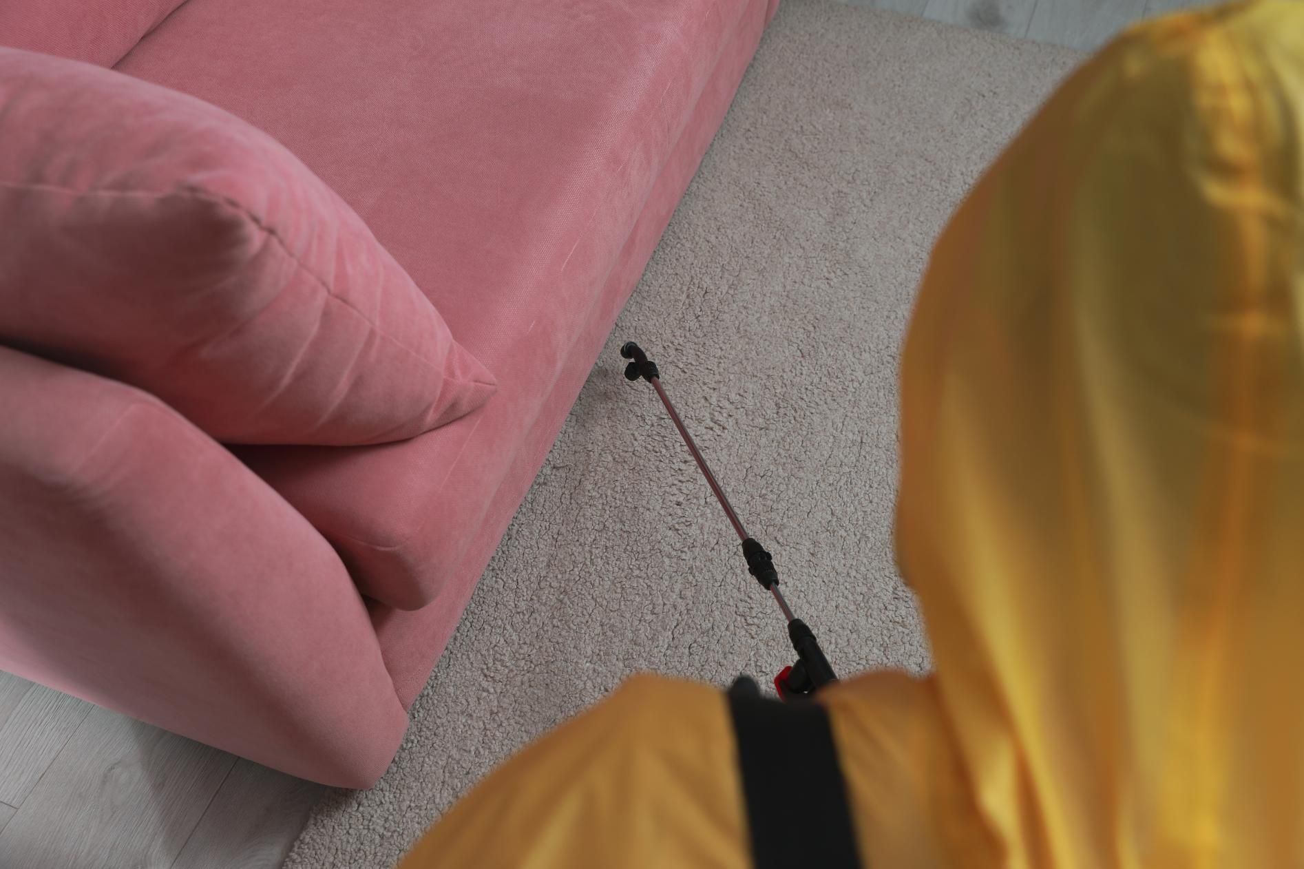 A Person in a Yellow Suit is Spraying a Pink Couch in a Living Room — Rozken Pest Control Services In Beechwood, NSW