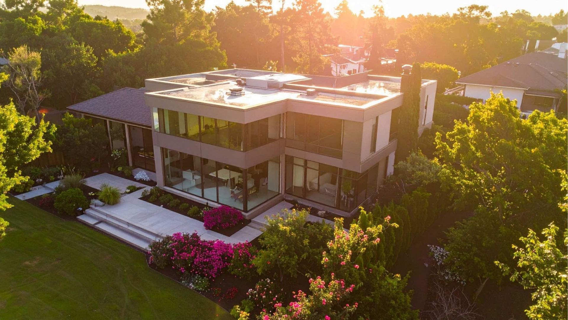 Modern two-story house with rooftop patio, surrounded by lush trees and pink flowers at sunset