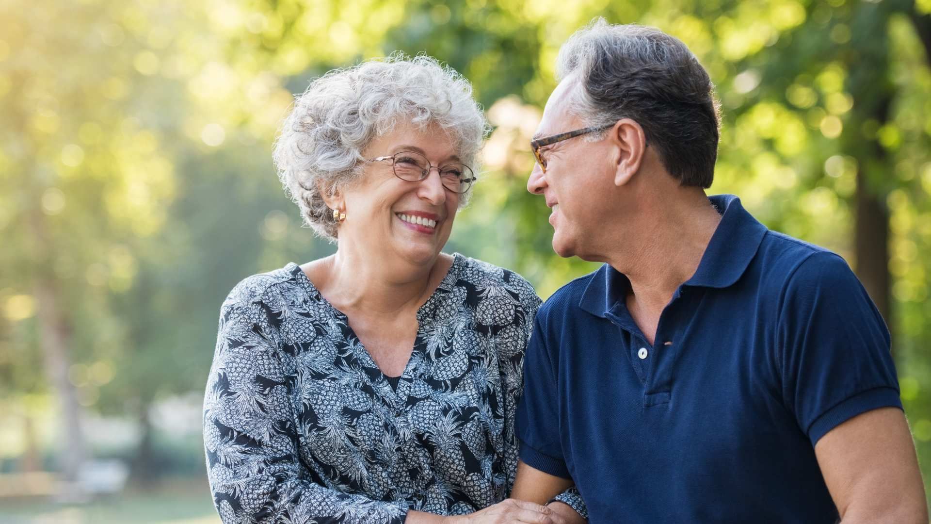 Two smiling people sit outdoors, looking at each other and laughing in a sunny park.