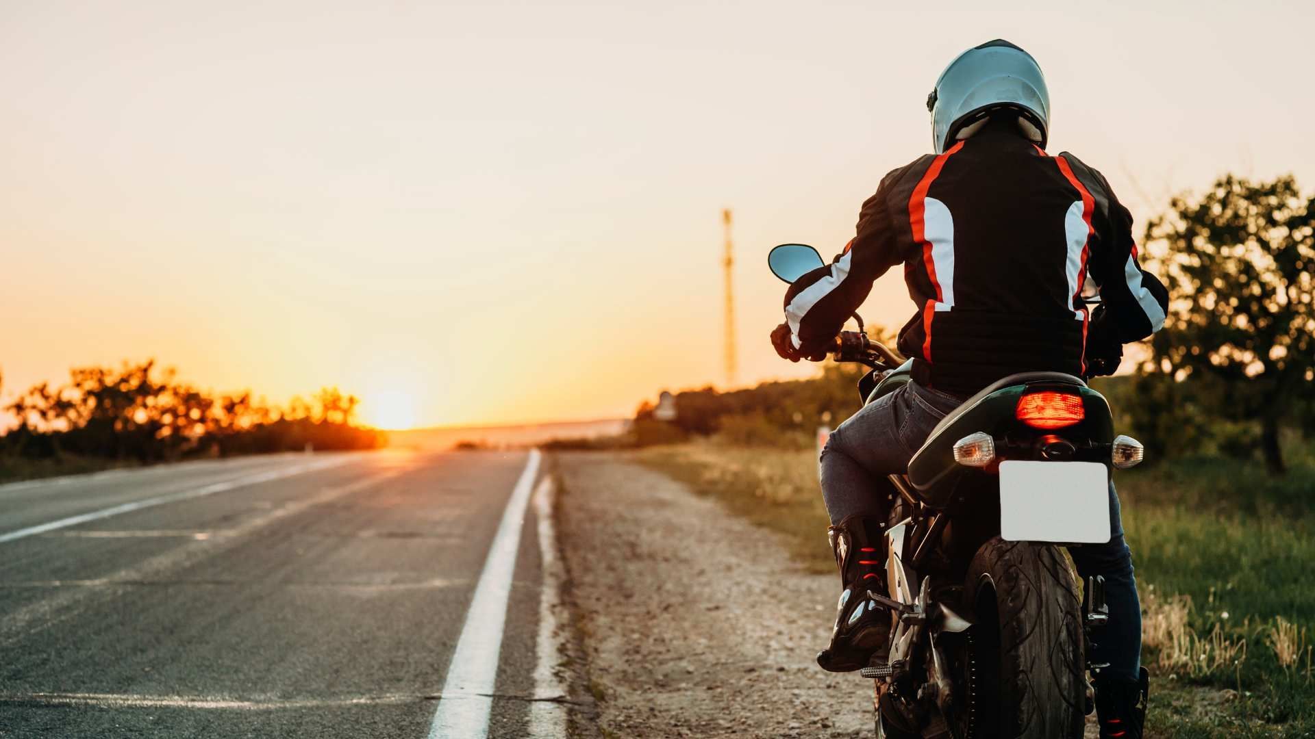 Motorcyclist riding along a road at sunset, viewed from behind.