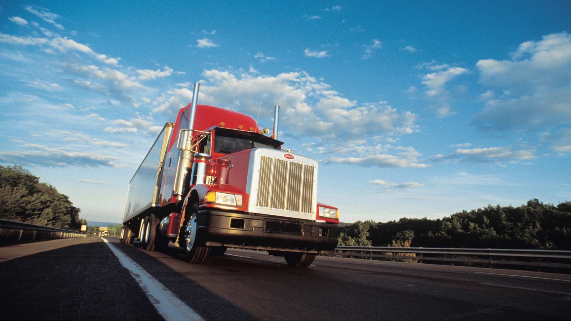 Red semi-truck driving on a highway under a blue sky with scattered clouds