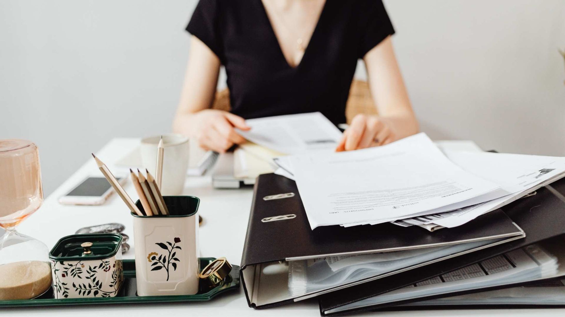 Person sorting papers at a desk with a laptop, notebook, mug, and pencil holder