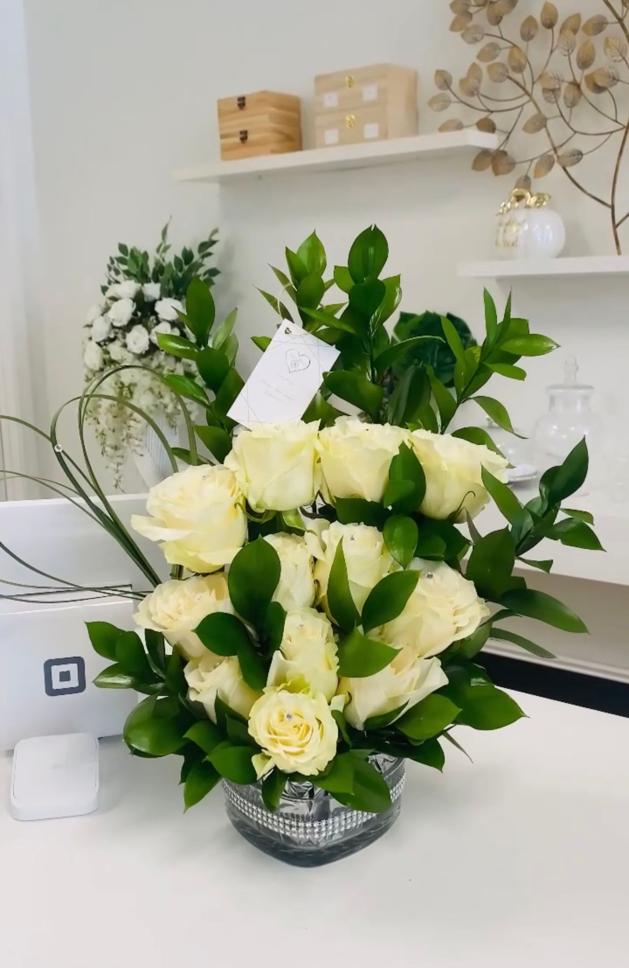 White roses and greenery arranged in a decorative vase on a white surface.