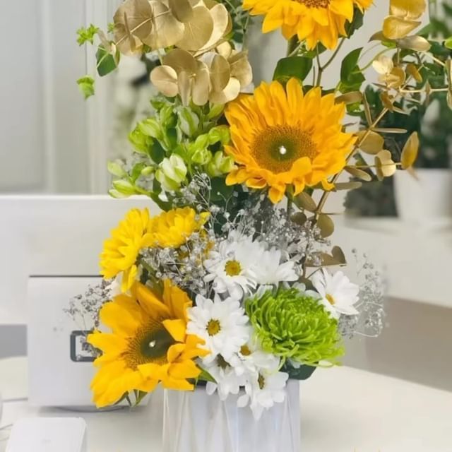 Vase with yellow sunflowers, white daisies, and green accents. Gold foliage in a bright room.
