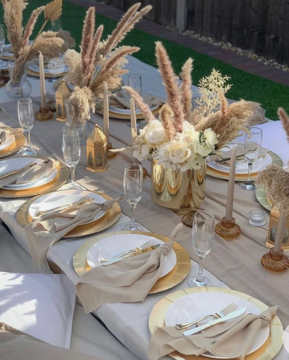 Elegant outdoor table setting with gold chargers, white plates, champagne flutes, and pampas grass centerpieces.